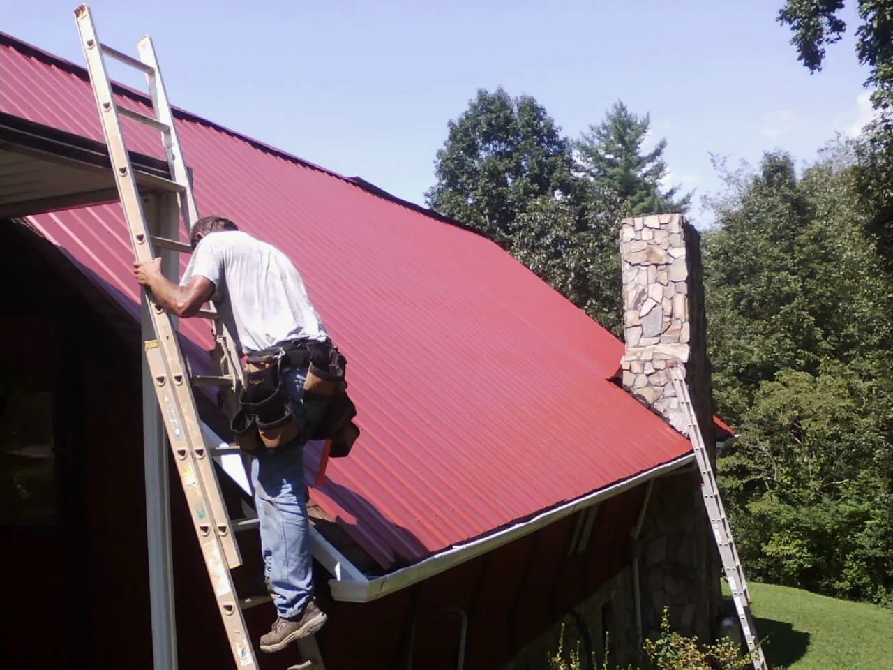 Roofer on a ladder beside a red metal roof and stone chimney — Soffit & Fascia Repair by Roofing Brothers in Angelus Oaks